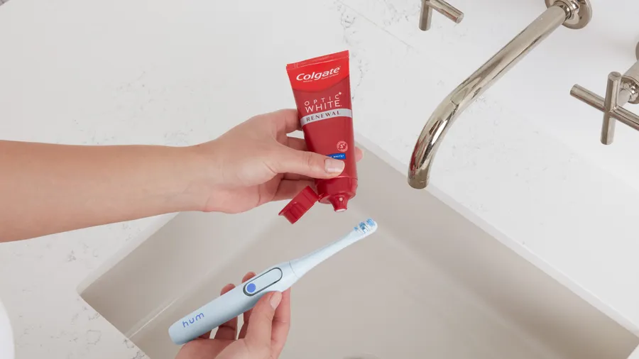 person applying colgate optic white toothpaste on blue colgate hum adult smart rechargeable electric toothbrush