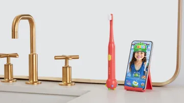 colgate hum smart battery powered coral red toothbrush for kids and a smartphone holder on the sink counter