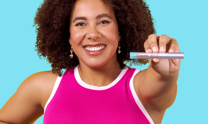 A smiling woman in a pink shirt holding a Colgate Optic White Express Teeth Whitening Pen.